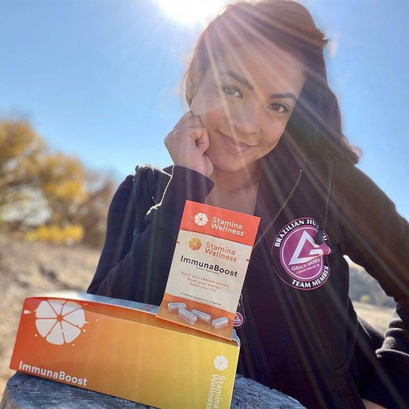 Woman smiling while leaning face on curled hand, her elbow leaning on a rock with a box, packet, and capsules of ImmunaBoost on the rock in front of her and the sun shining over a desert landscape behind her