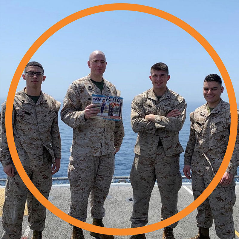 Four U.S. Marines smiling while standing on the deck of a ship at sea, one of them holding up a calendar, an orange ring stylistically placed over the entire scene