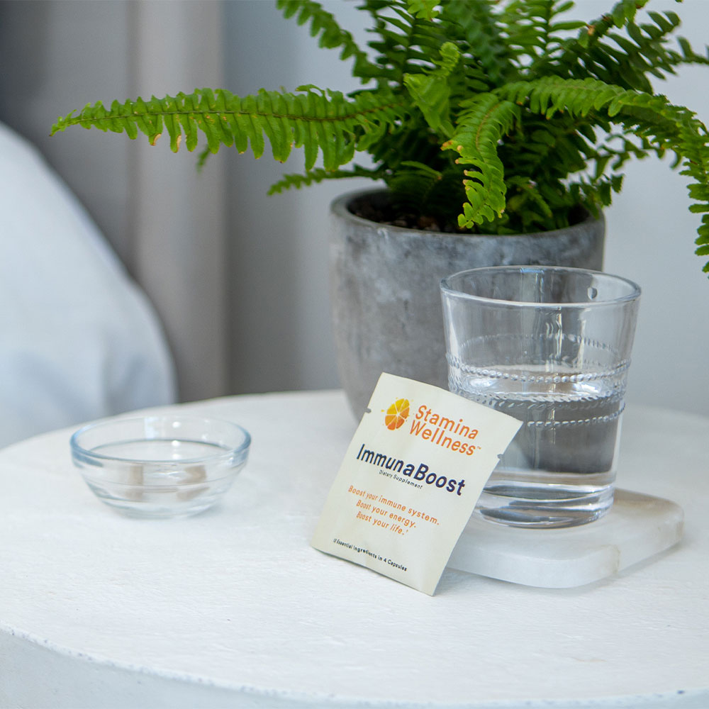 A packet of ImmunaBoost leaning up against a glass of water on a white marble coaster sitting on a marble nightstand table with a small glass bowl and green plant resting atop and a bed in the background